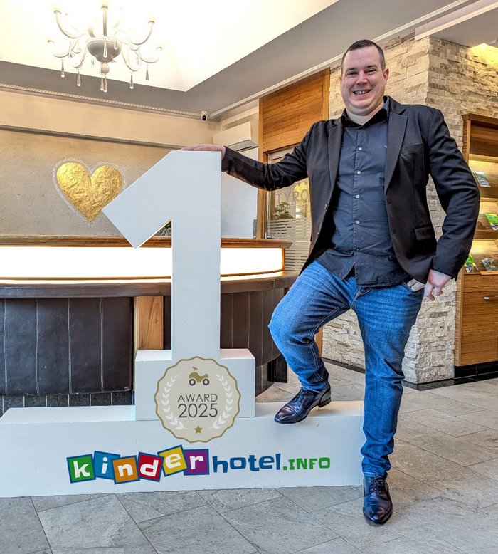 Mann steht an der Rezeption neben einem großen 1st-Place-Schild für kinderhotel.info Award 2025