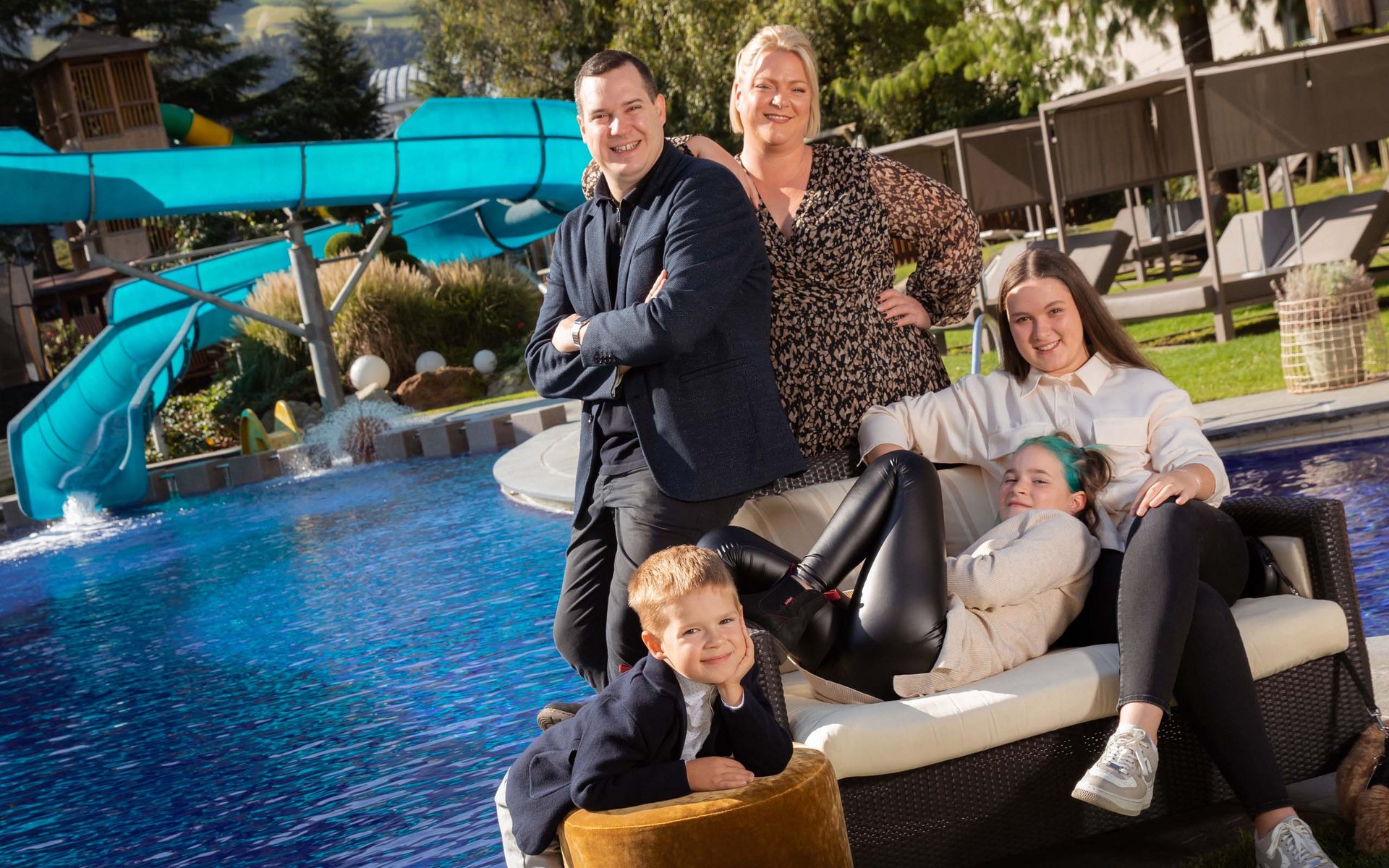 Familie posiert entspannt neben Pool und blauer Wasserrutsche im Sonnenschein