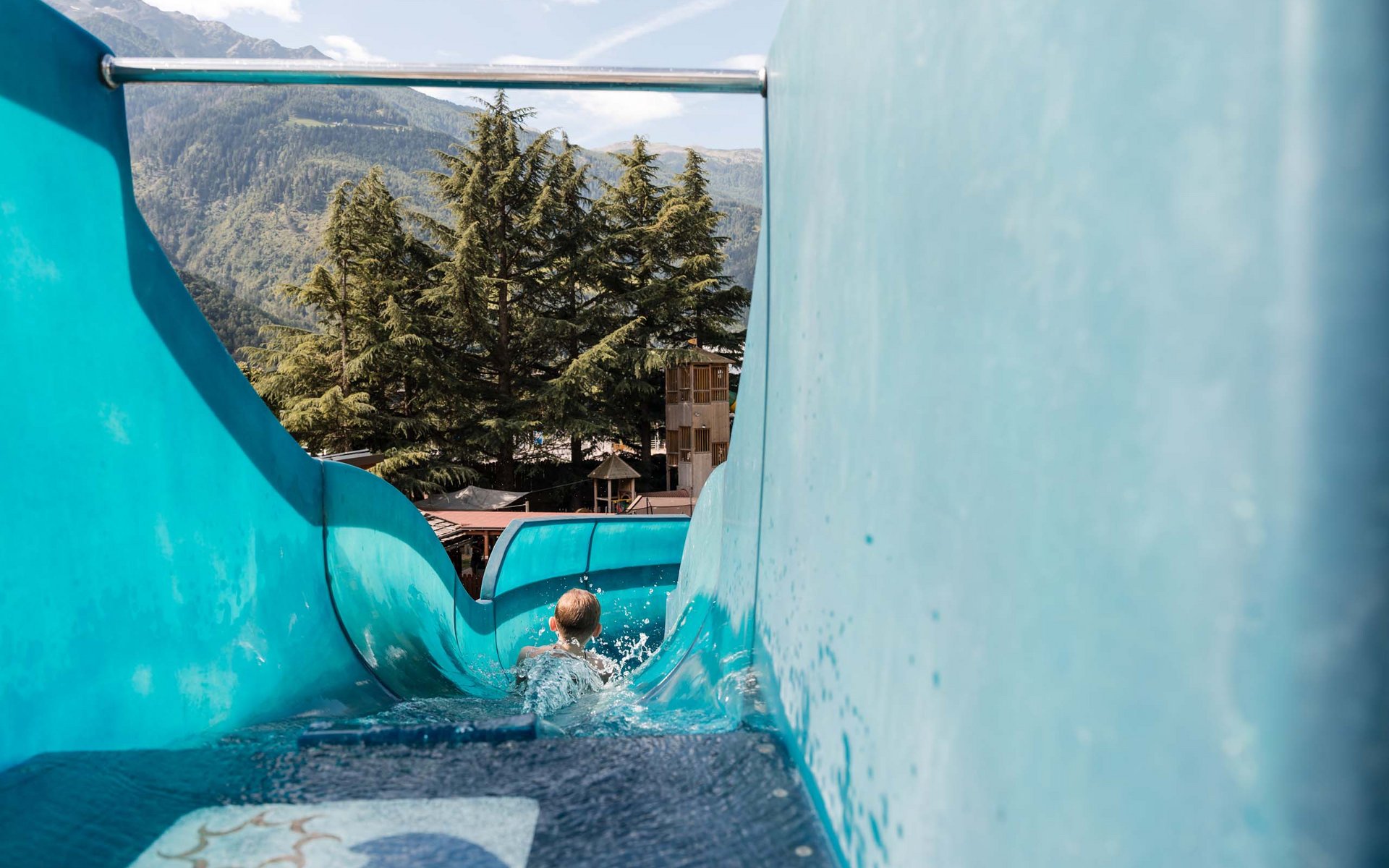 Kind rutscht auf einer blauen Wasserrutsche mit Bergen und Bäumen im Hintergrund