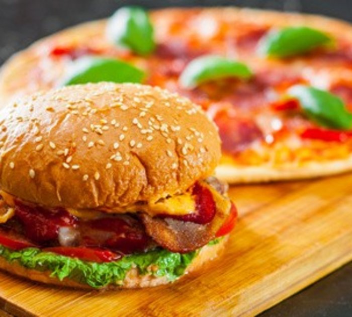 Burger with lettuce and bacon in front of a pizza with basil on a wooden board