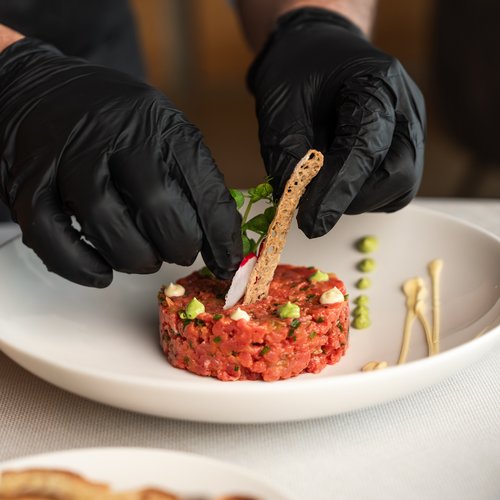 Person mit schwarzen Handschuhen arrangiert Tatar auf weißem Teller