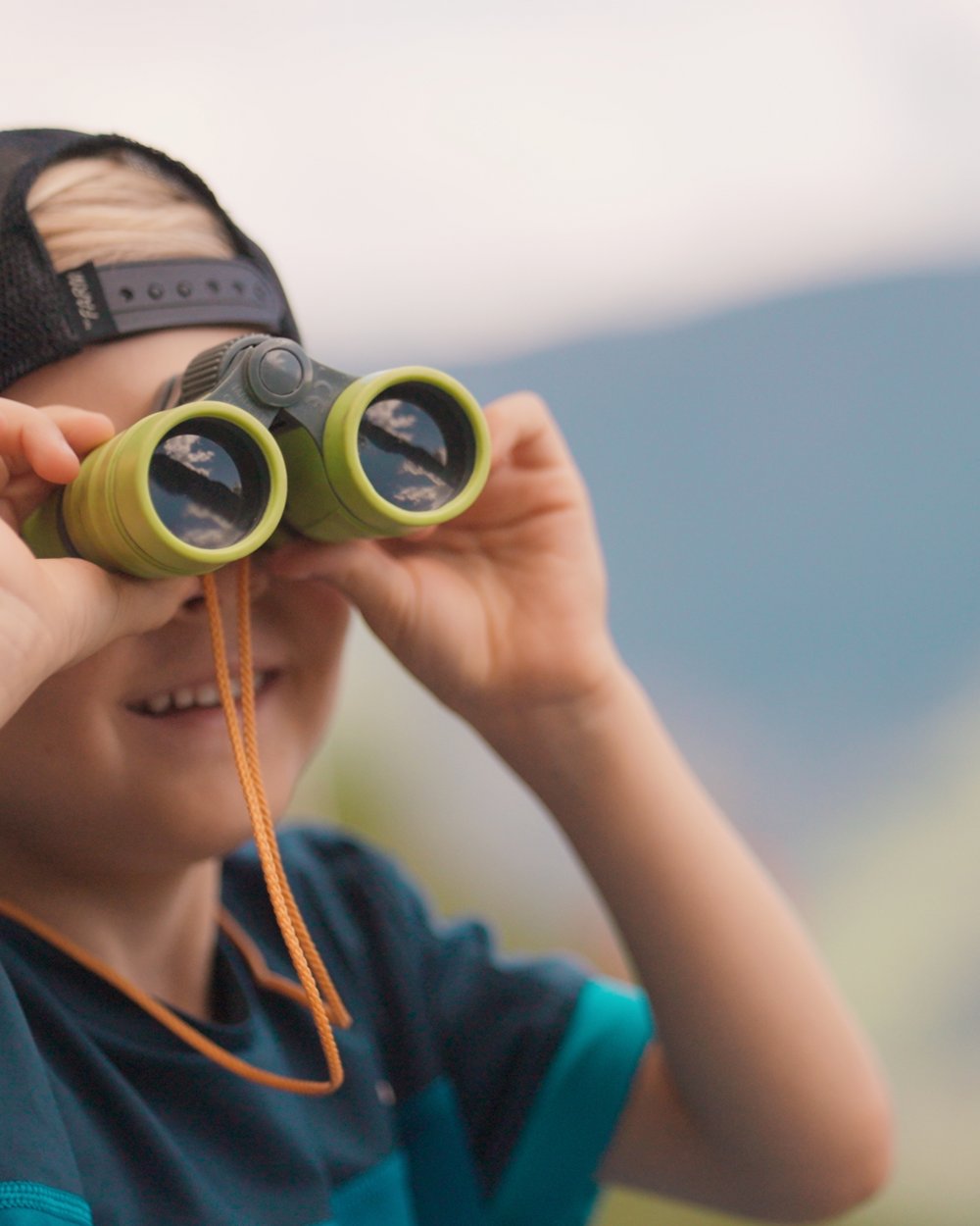 Spiel und Spaß im Hotel mit Kinderbetreuung in Südtirol Junge mit schwarzer Kappe schaut durch grüne Ferngläser im Freien