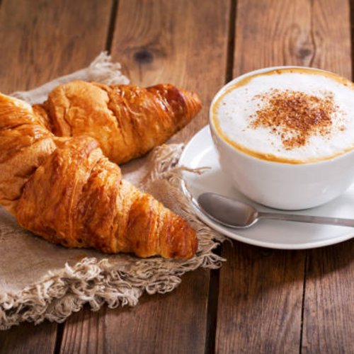 Two croissants on cloth next to a cappuccino cup on wooden table