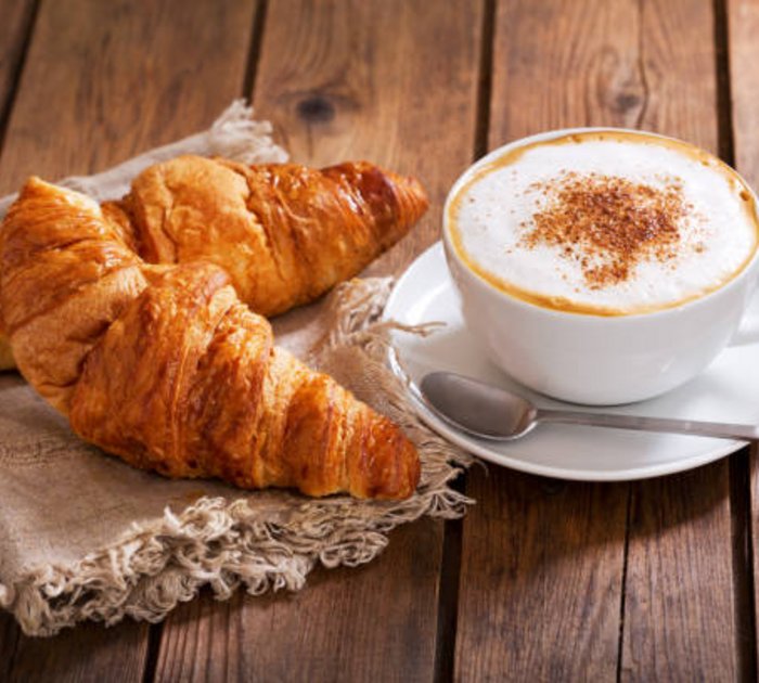 Two croissants on cloth next to a cappuccino cup on wooden table