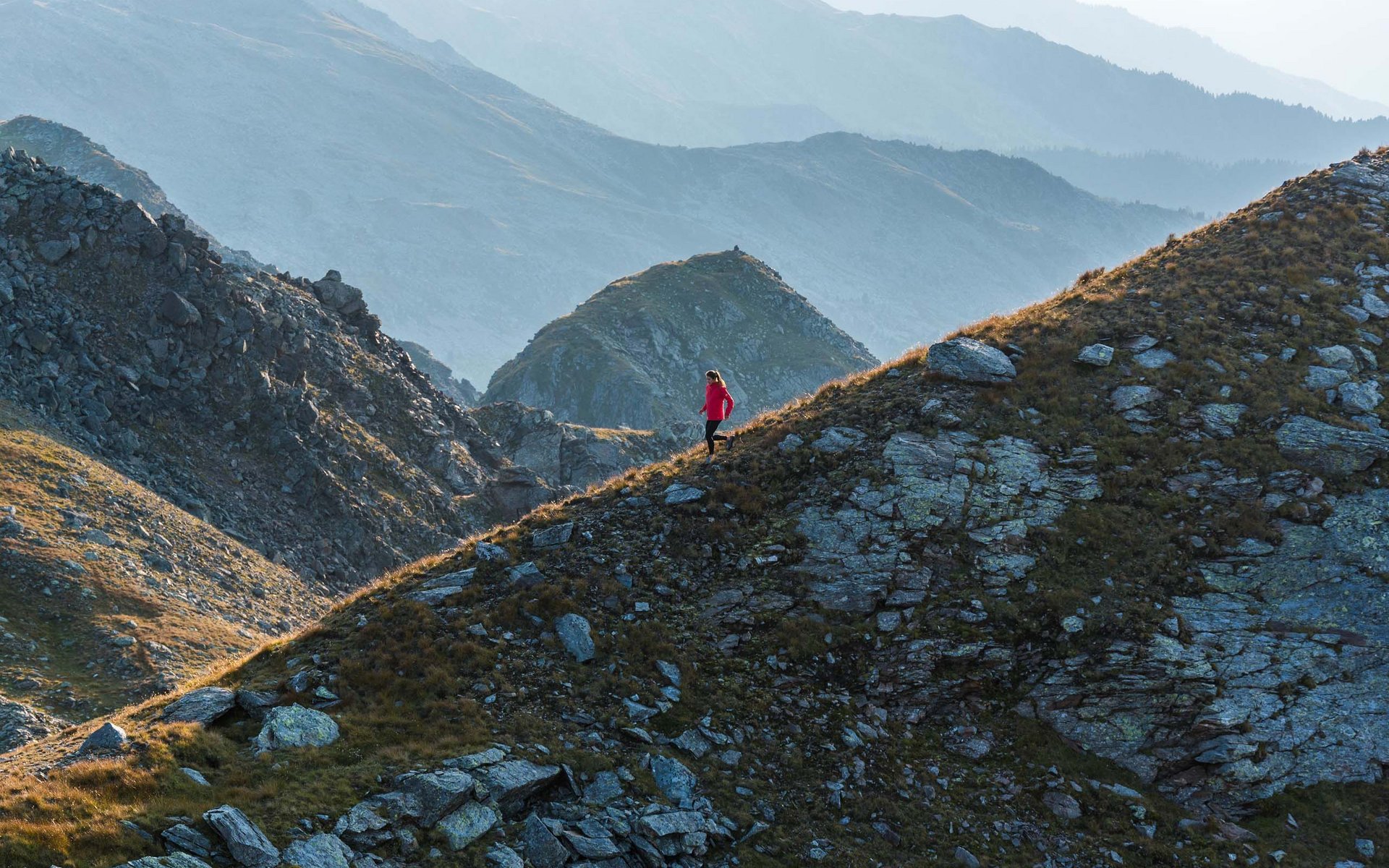 Wanderin in roter Jacke auf felsigem Bergpfad bei Sonnenlicht