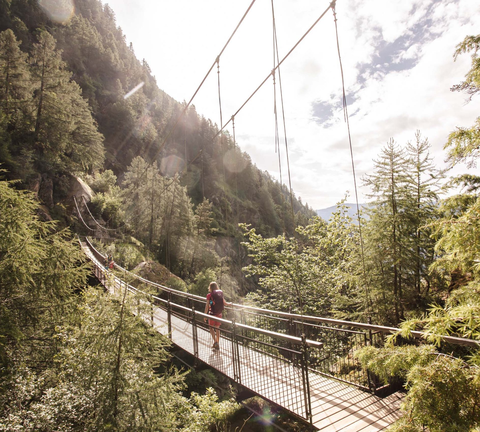 Person walking on a suspension bridge in a sunny forested mountain valley