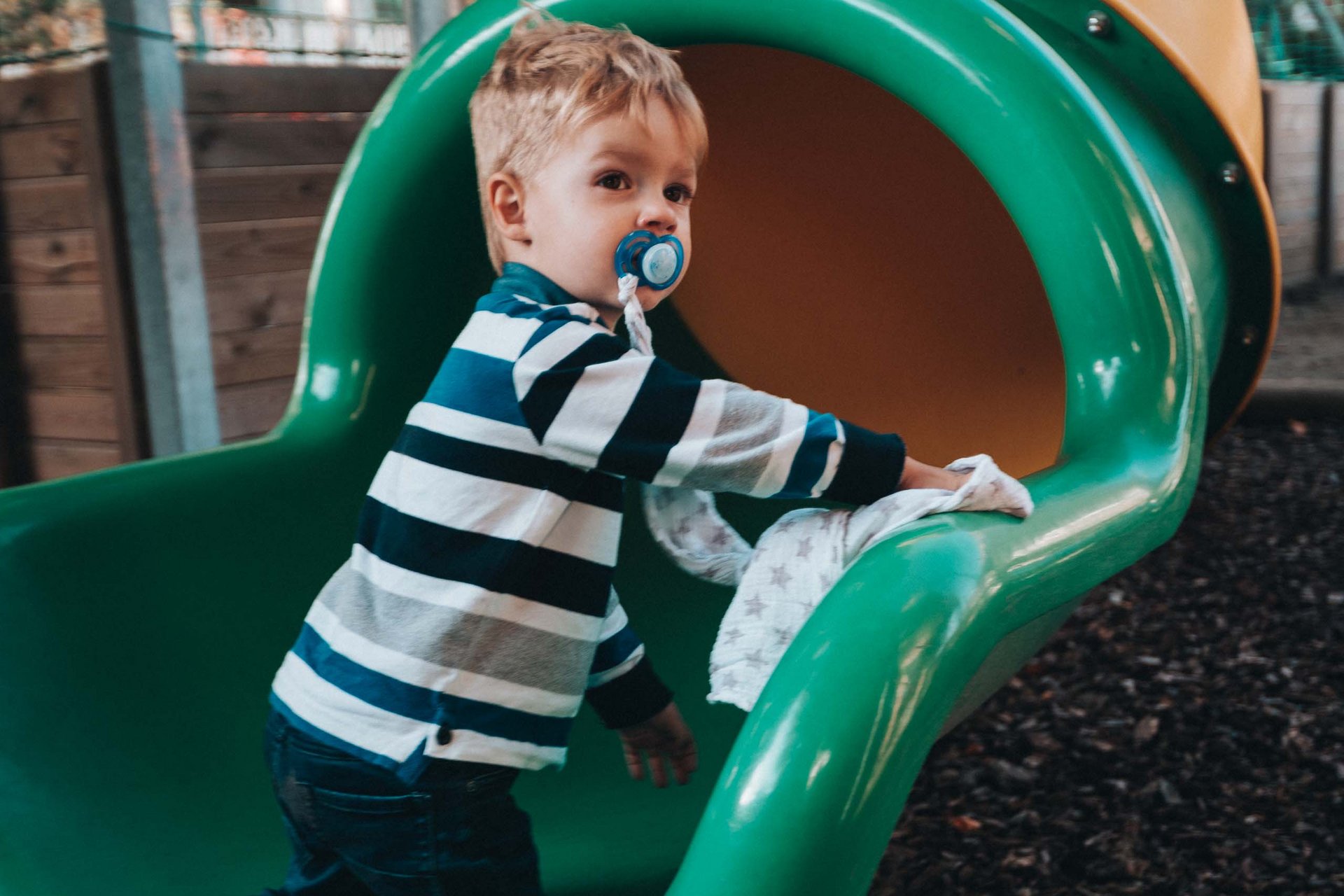 Bambino con ciuccio che si arrampica sullo scivolo verde al parco giochi