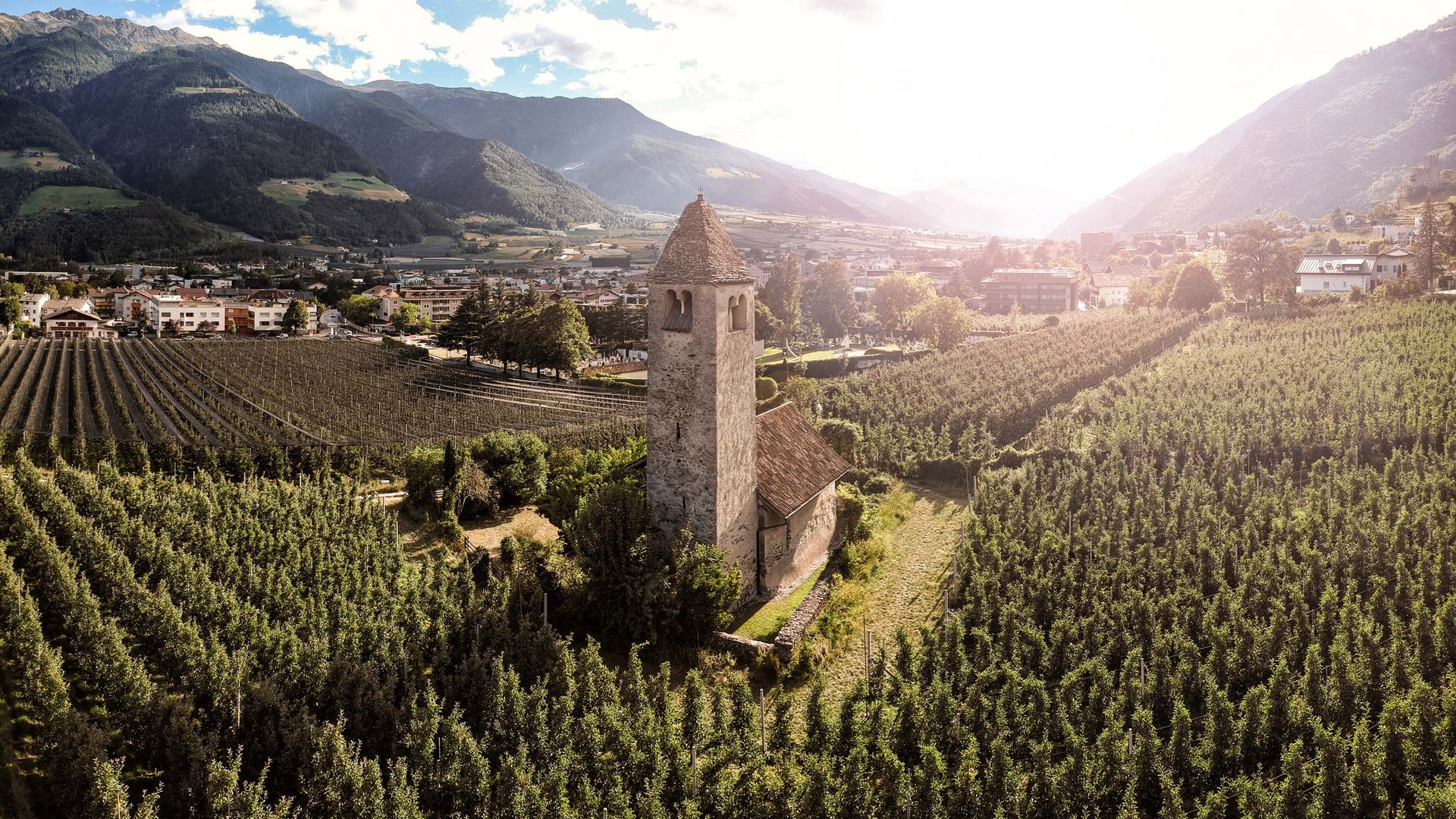 Historischer Kirchturm inmitten von Apfelplantagen mit Bergen im Hintergrund